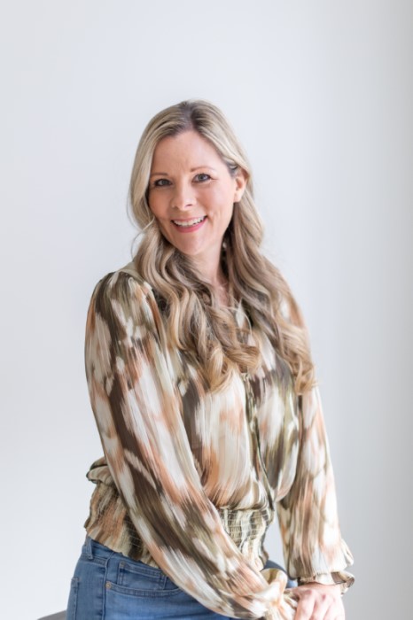 A professional headshot of Pizzajules, a blonde woman with her hair curled sitting in front of a gray background.